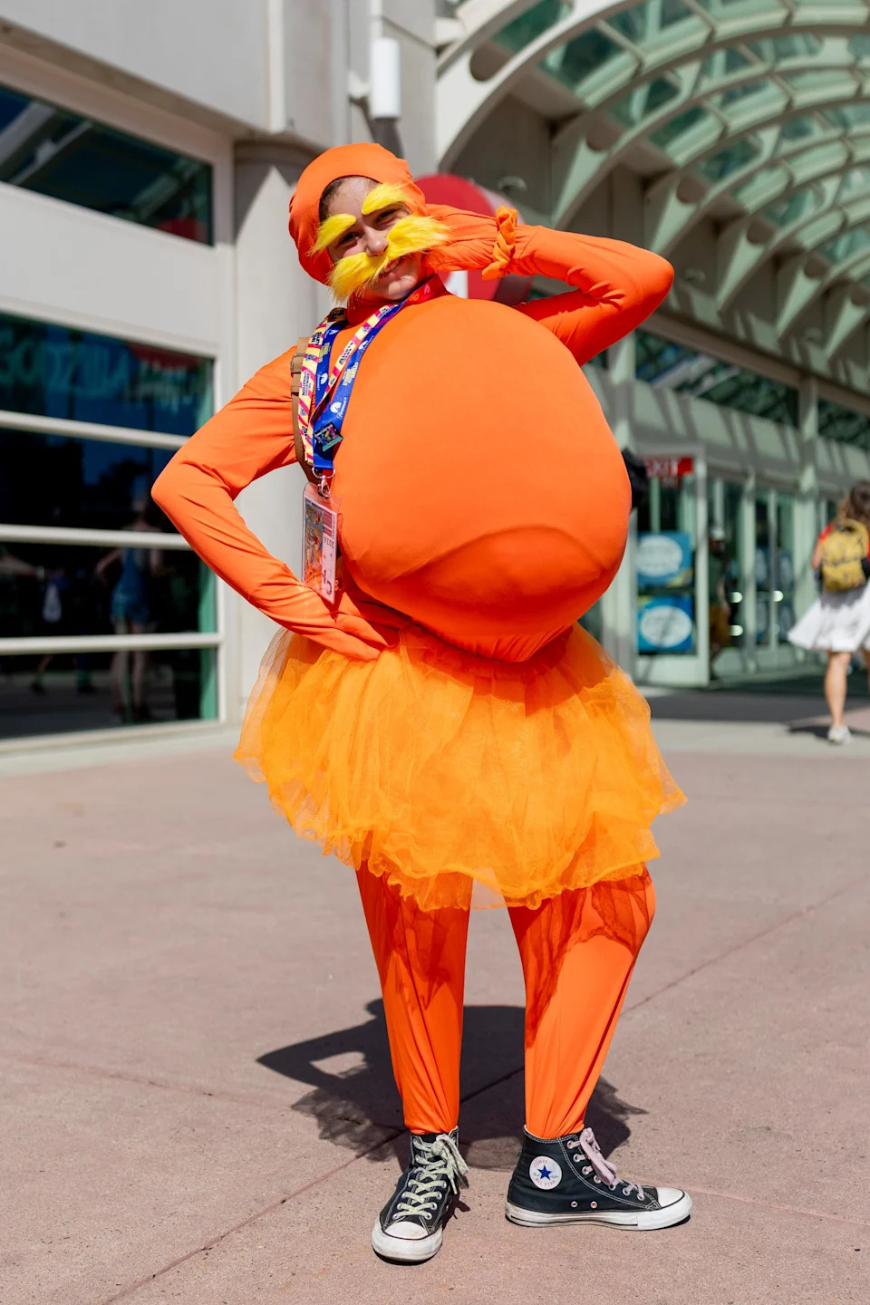 Person in an orange costume with a tutu, moustache, and sneakers poses playfully in a convention setting