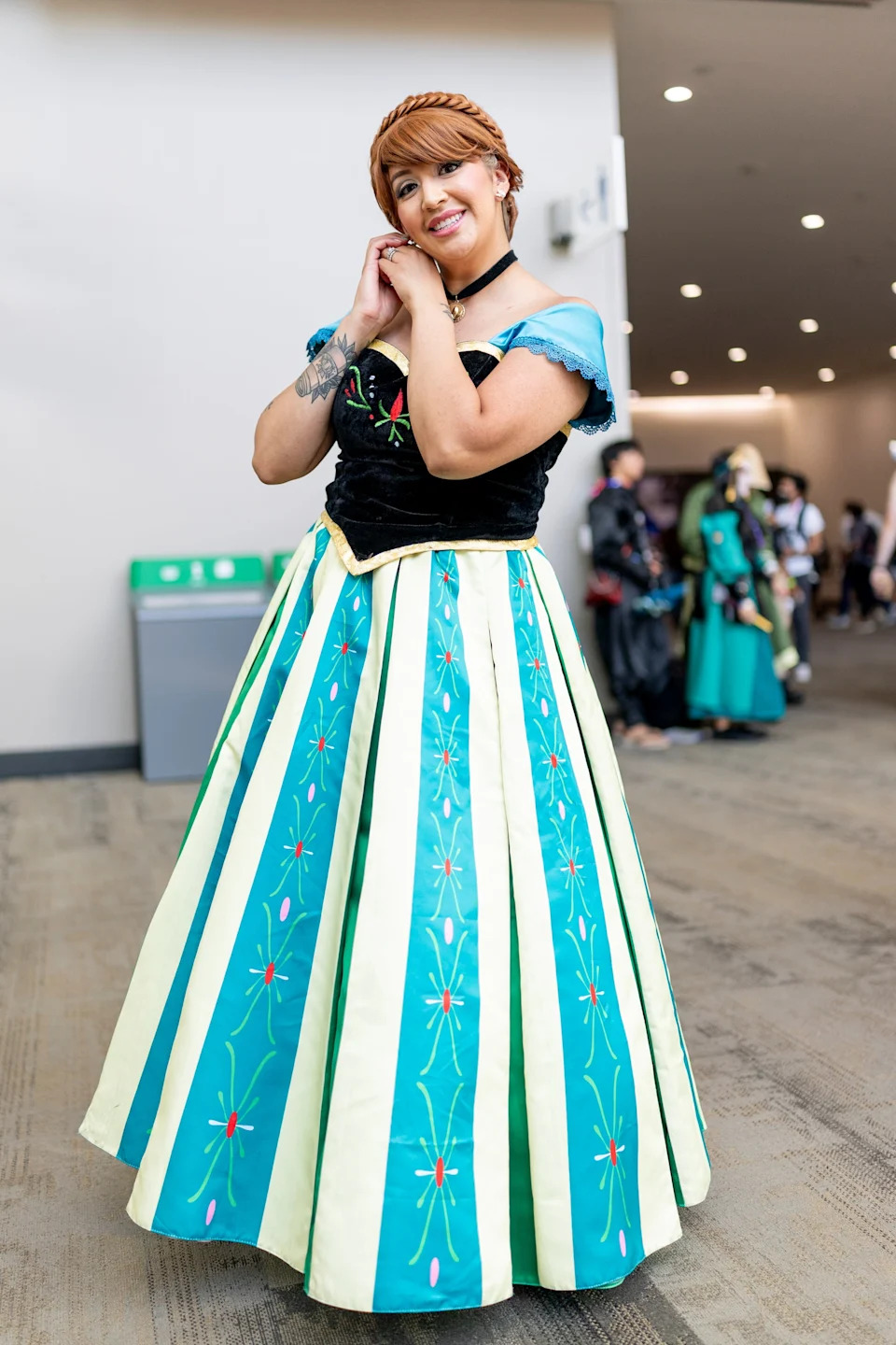 Person in princess-style costume with embroidered floral patterns stands in a convention setting, smiling with hands clasped near the face