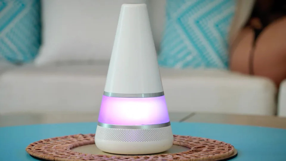 A cone-shaped air freshener with a glowing purple band sits on a woven mat on a table, with patterned pillows in the background