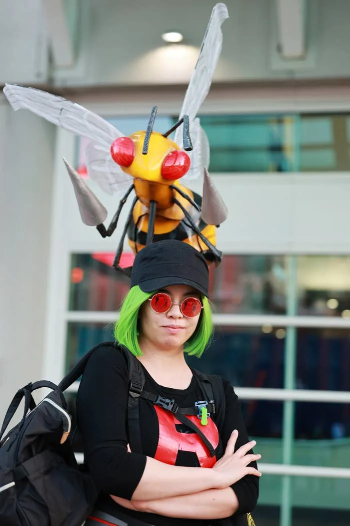Person in costume with a large insect headpiece, wearing sunglasses, a backpack, and a cap. They're standing confidently with arms crossed
