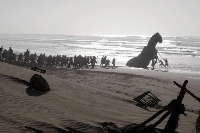 A group of people pulling a large horse statue across a beach, with waves in the background