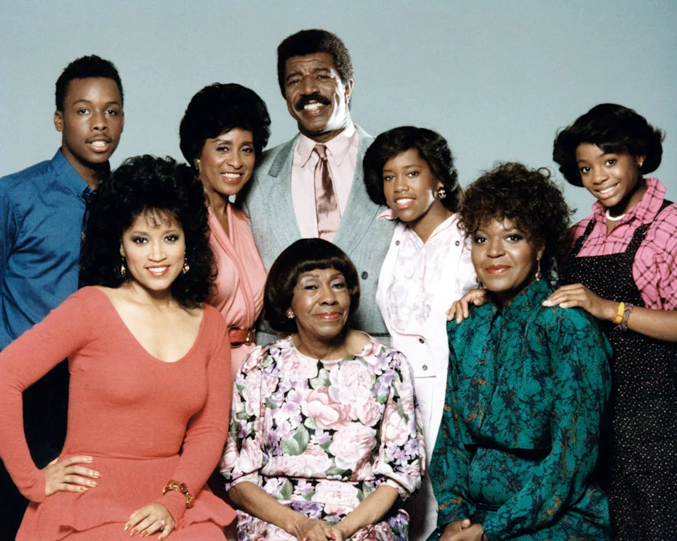 A group photo of eight people smiling, featuring various fashion styles from the era. Central focus is on the person in floral attire seated in front