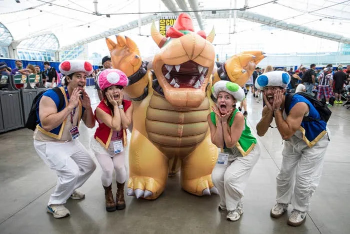 Four people in Toad costumes pose excitedly next to a large inflatable Bowser character at a comic convention