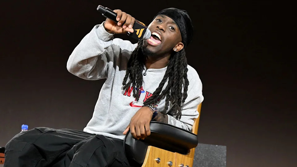 Kai Cenat with long hair is enthusiastically speaking into a microphone while sitting on a chair, wearing a gray sweatshirt and black pants.