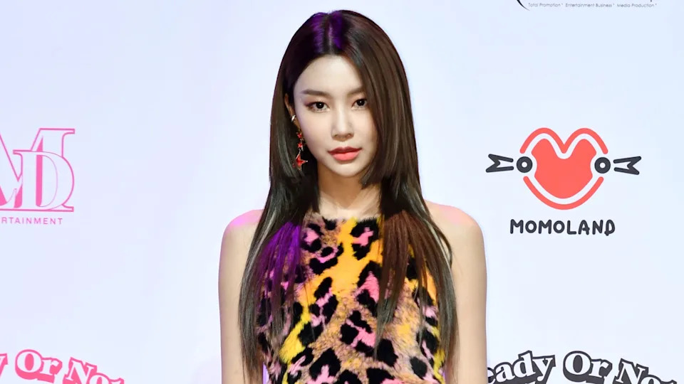 K-pop singer Hyebin in a colorful leopard-print dress poses in front of a backdrop with "MOMOLAND" and other logos.