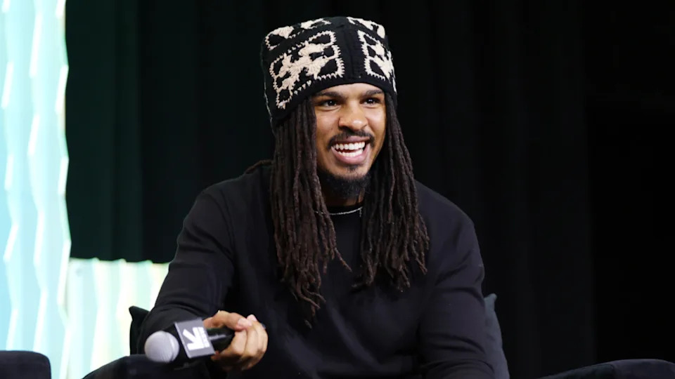 Keith Lee with long dreadlocks, wearing a patterned hat, smiles while holding a microphone on stage.