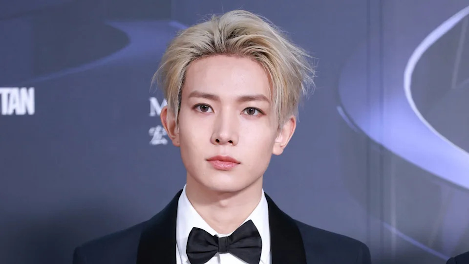 K-pop star Heeseung with blonde hair in a tuxedo and bow tie at a formal event, standing against a dark background.
