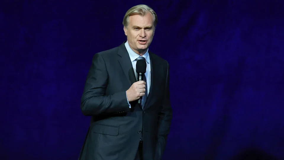 Christopher Nolan in a suit holding a microphone stands against a dark blue background, speaking.