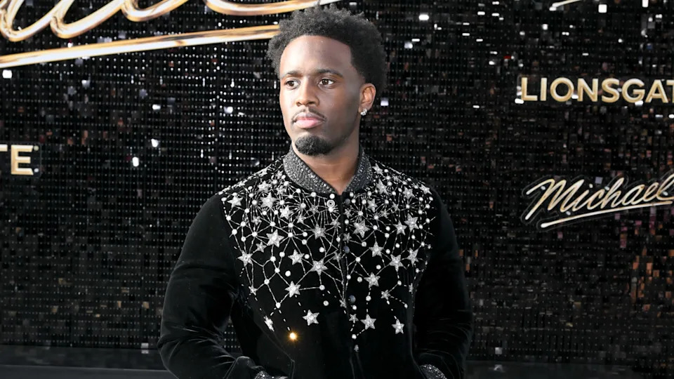 Kai Cenat in a black jacket with star patterns stands in front of a sparkly backdrop with "Lionsgate" and "Michael" visible.