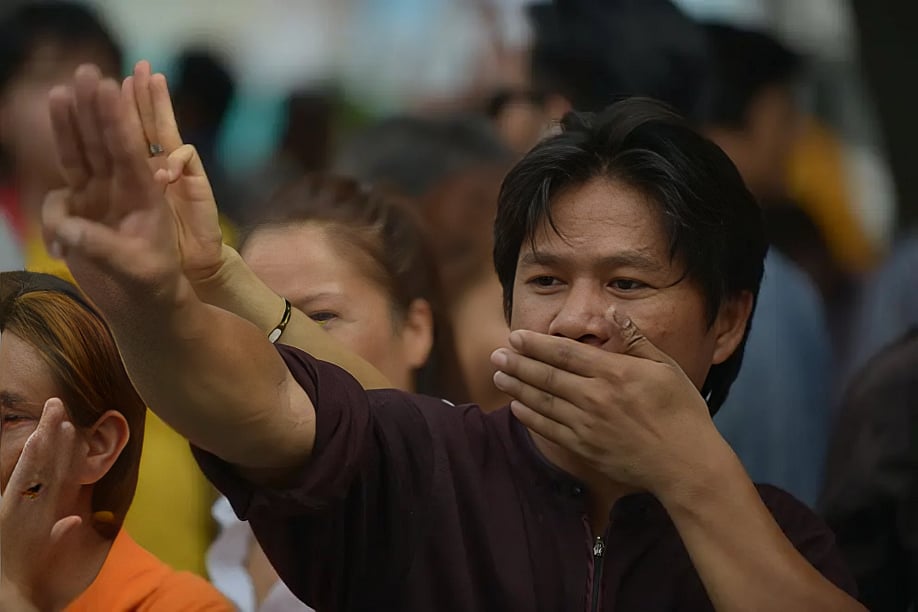 Close-up shot of a man covering his mouth with his right hand while extending his left arm forward in a three-finger salute. He is standing in a dense crowd of demonstrators who are also raising their hands in pro-democracy protest gestures.