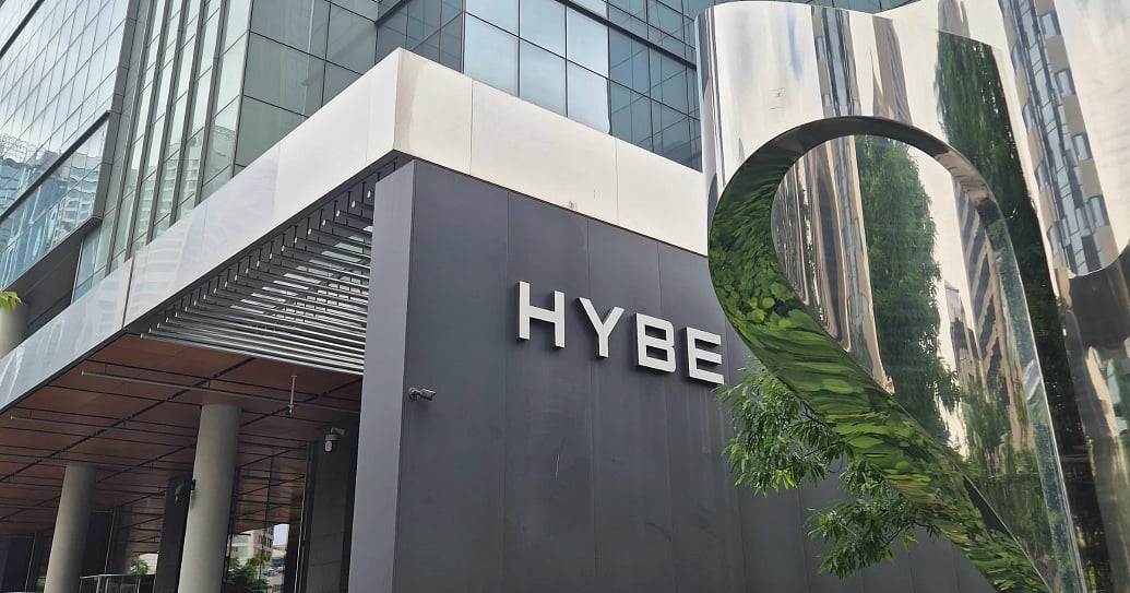 A modern exterior view of the K-pop giant, HYBE's headquarters in Seoul, South Korea. The company logo is mounted on a dark facade next to a polished, curved chrome art installation that reflects surrounding green trees.