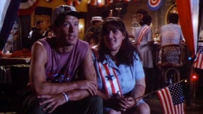 Brian Wimmer as Boonie and Ricki Lake as Holly, sitting next to each other surrounded by flags and independence day decor in China Beach.