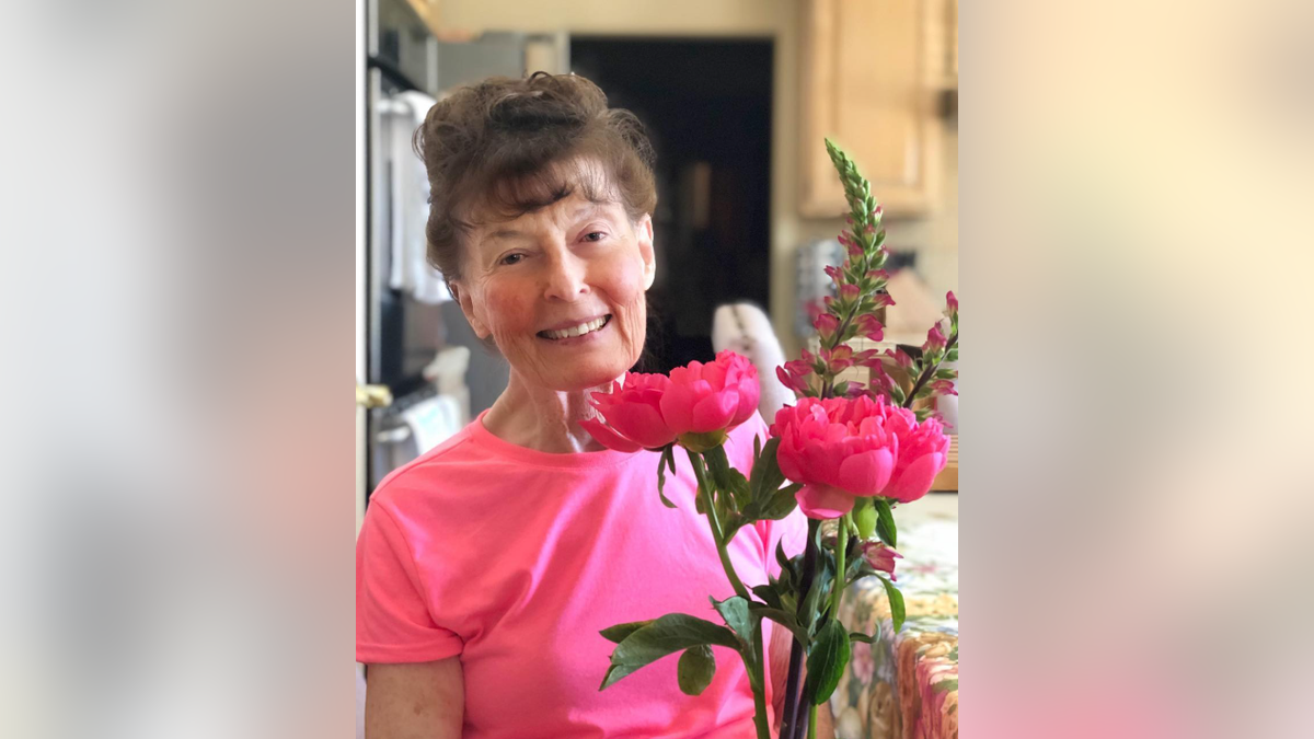 Linda Perhacs smiles while seated indoors holding a bouquet of vibrant pink flowers.