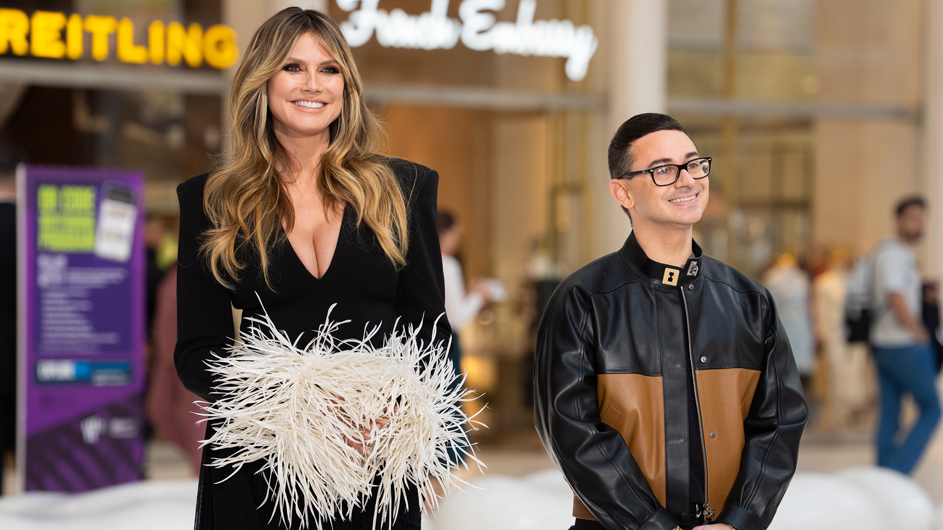 A promotional image for Project Runway featuring host Heidi Klum in a low-cut black gown with feathered cuffs alongside mentor Christian Siriano in a black and brown leather jacket, both smiling for an outdoor event.