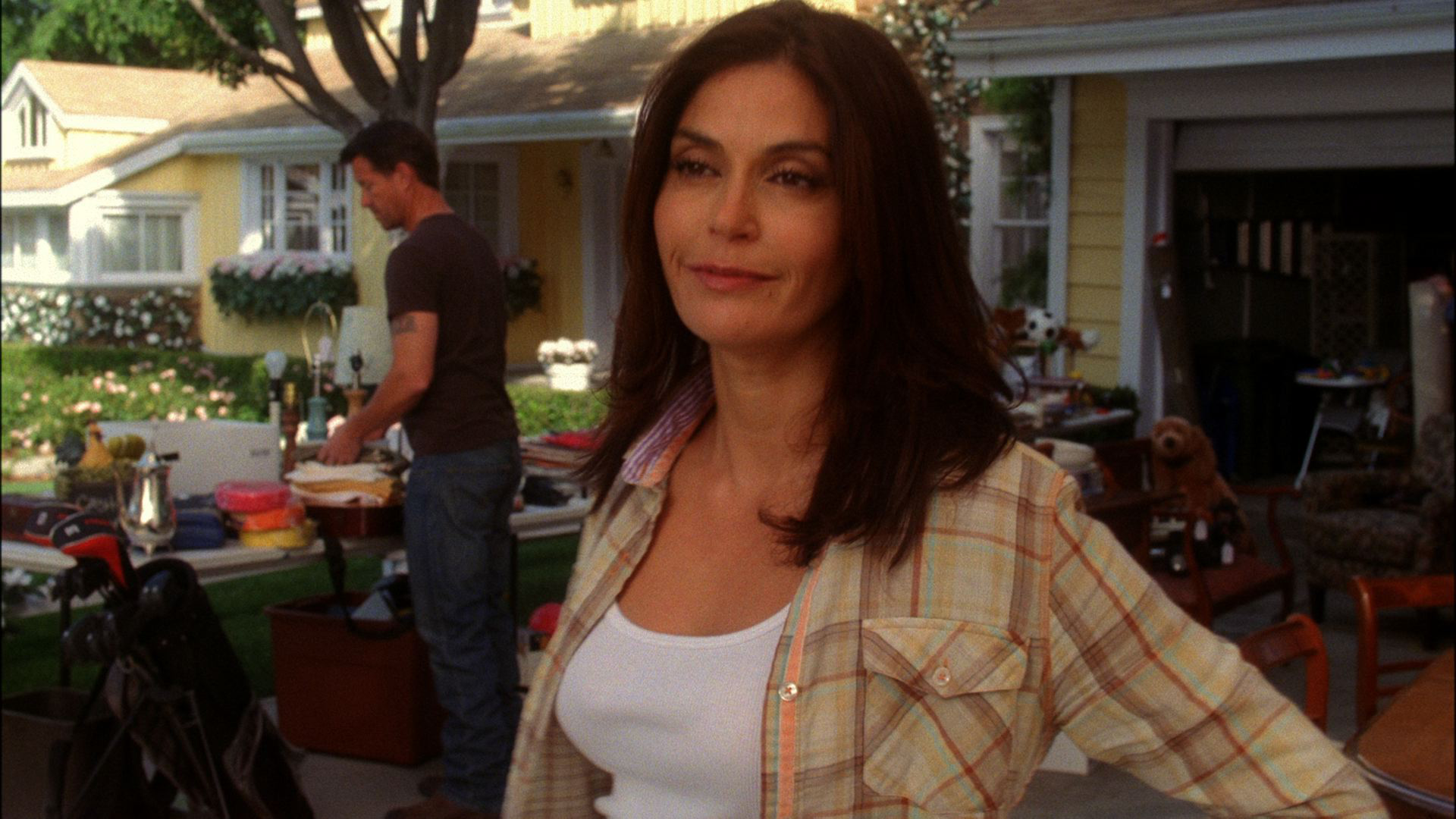 A scene from "Desperate Housewives" featuring Susan standing outdoors in a plaid shirt over a white tank top, smiling slightly while Mike works in the background near tables of items at what appears to be a yard sale.