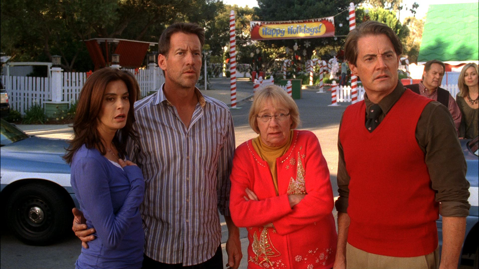 A scene from "Desperate Housewives" featuring Susan, Mike, Karen McCluskey, and Orson standing outside on Wisteria Lane during a holiday event, looking forward with shocked and worried expressions.