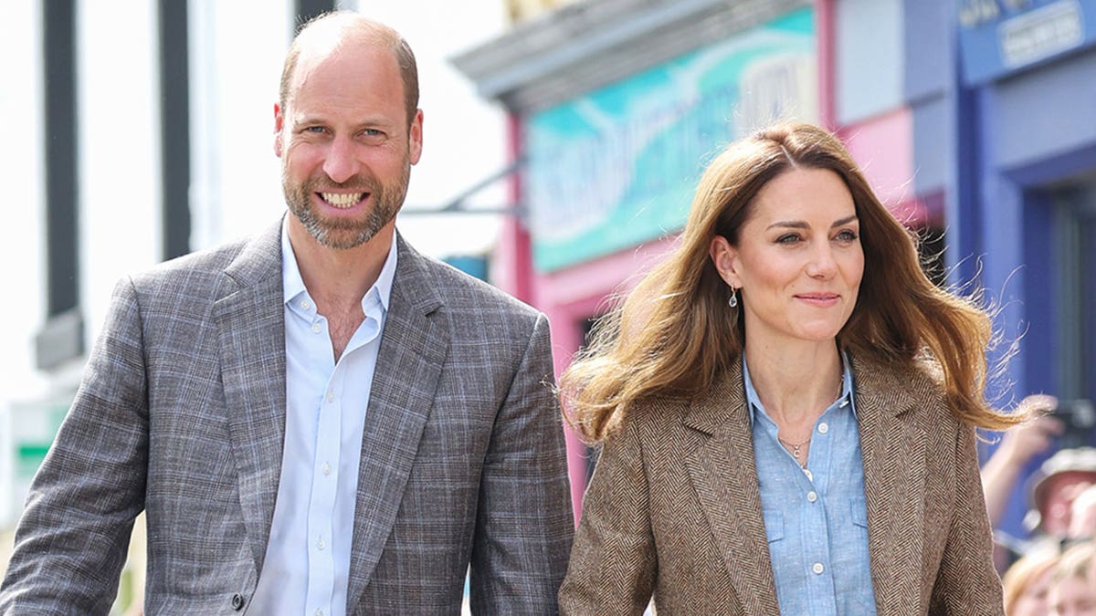 Prince William and Kate Middleton smiling as they walk side-by-side in matching blazer ensembles carrying out royal duties in the U.K.