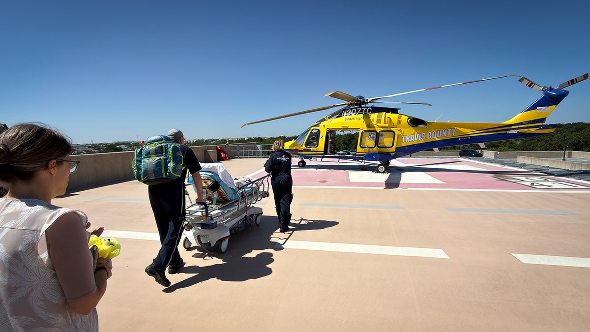 Medical personnel push a patient on a stretcher toward a helicopter on a helipad.