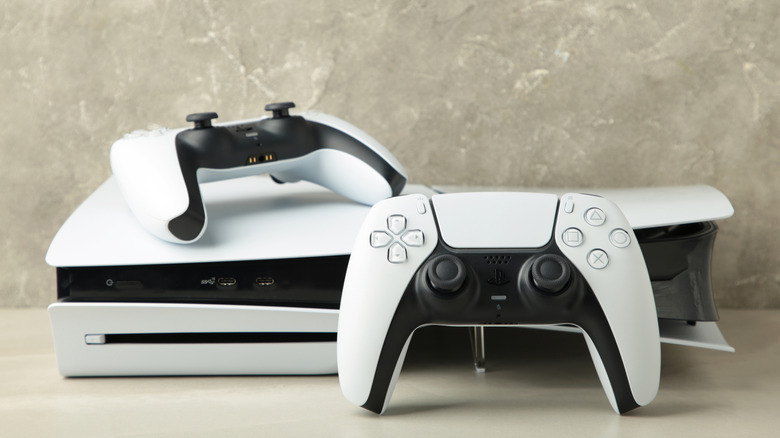 A PlayStation 5 and two DualSense controllers in front of a stone-textured wall