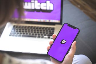 A person holds a smartphone displaying the Twitch app while a laptop in the background shows a Twitch screen.