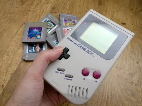 Hand holding Nintendo Game Boy. With consoles to the right.