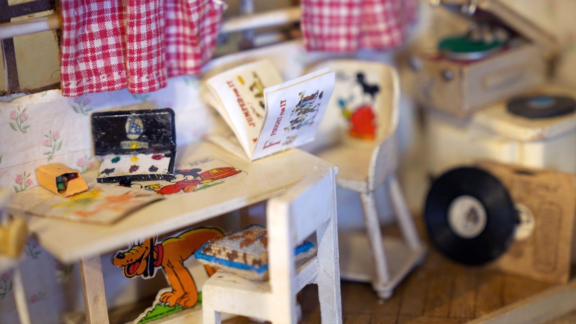 Another dolls house which is more modern with white chair and table, on which sits a miniature set of paints and colouring pens with disney characters coloured in around it. in the background there is a record player with miniature vinyl record discs dotted around.
