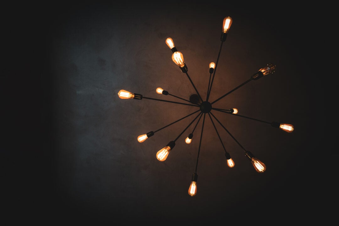 A ceiling light fixture designed with multiple exposed bulbs radiating outwards on metal arms, resembling a starburst pattern, set against a dark ceiling. The bulbs emit a warm glow, creating a cozy atmosphere.