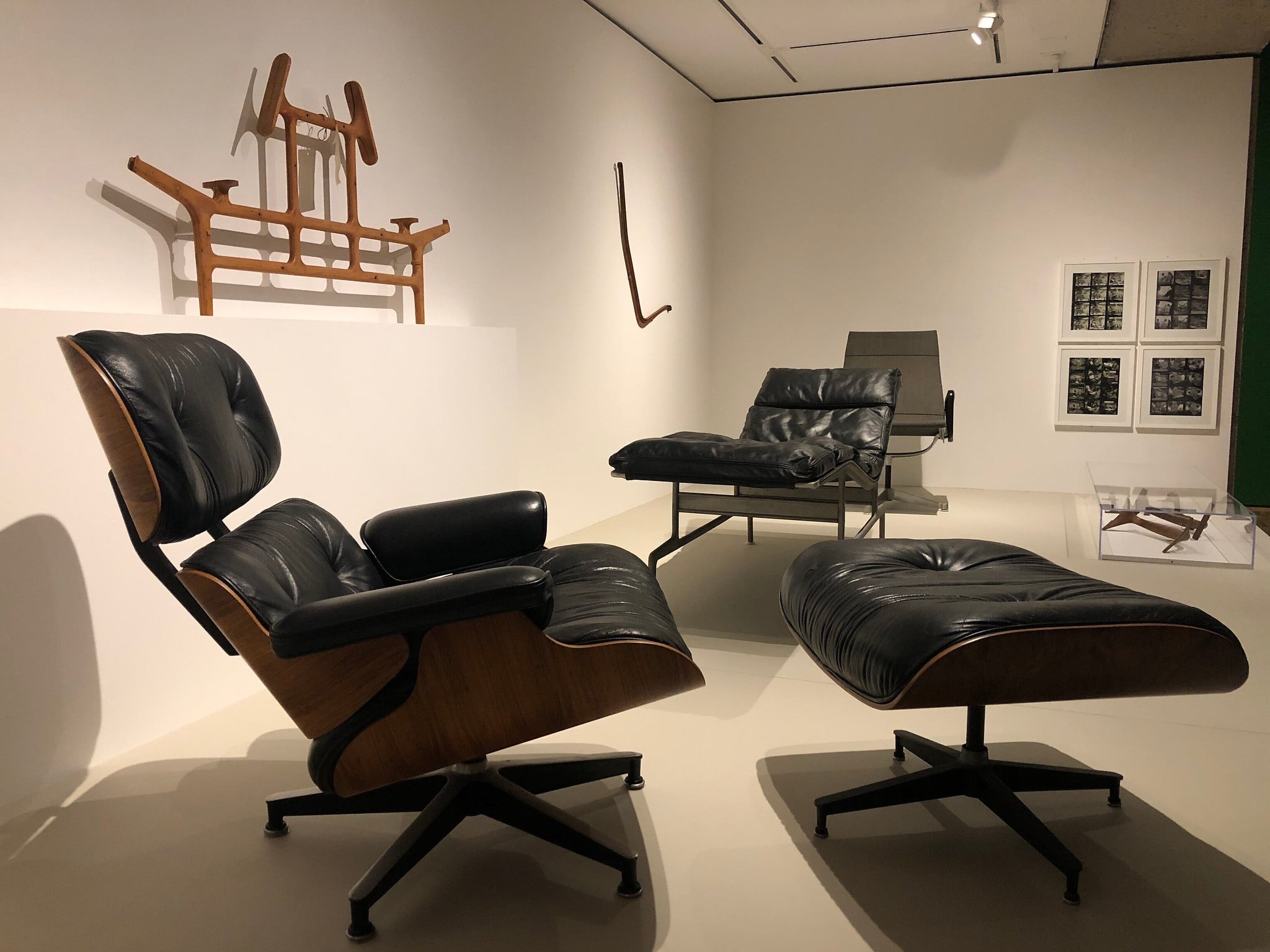 A gallery room featuring modern furniture, including a black leather and wood lounge chair with an ottoman, another sleek black chair, and wooden frames against white walls. Minimalist design and geometric art pieces are displayed in the background.