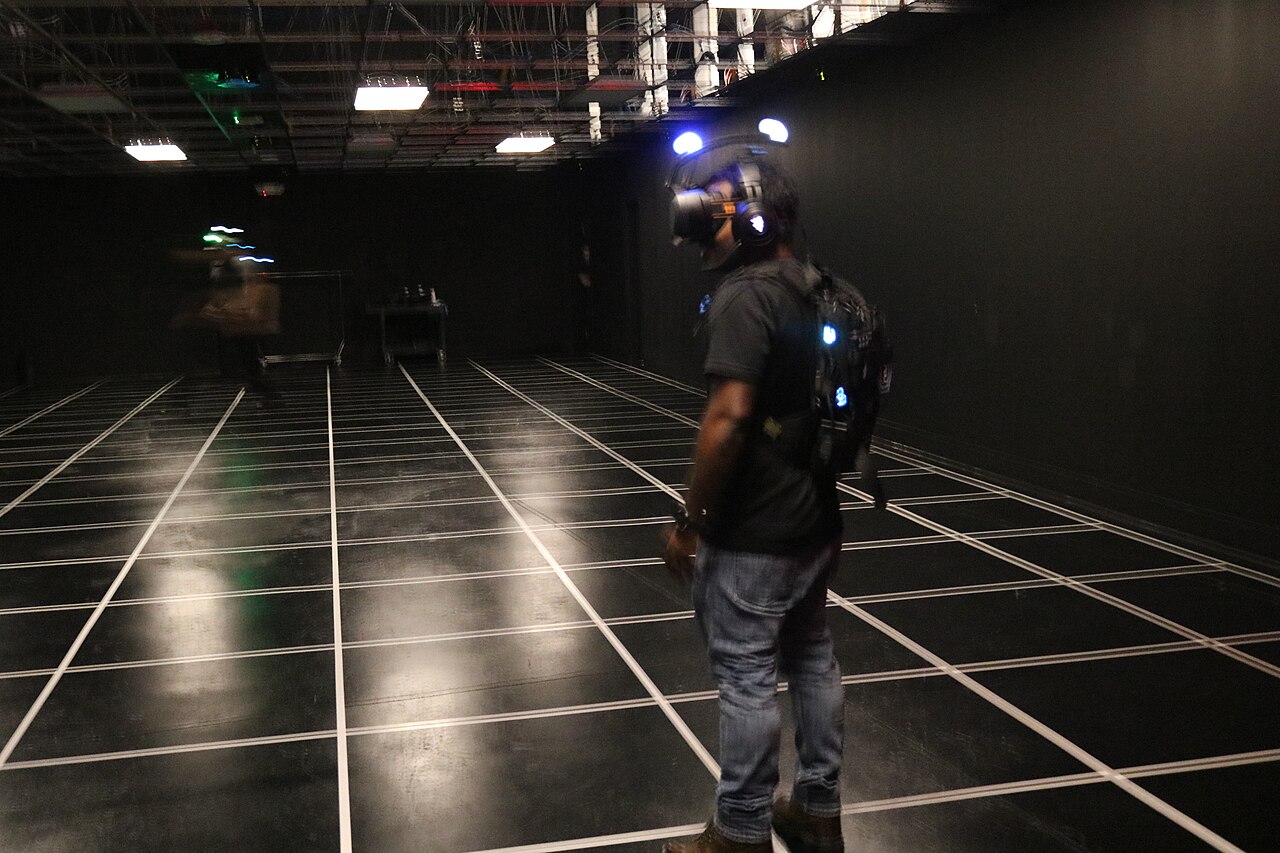 A player standing alone in a darkened warehouse arena with a white grid painted across the floor, wearing a VR headset with blue tracking lights and a backpack computer, another player blurred in the distance