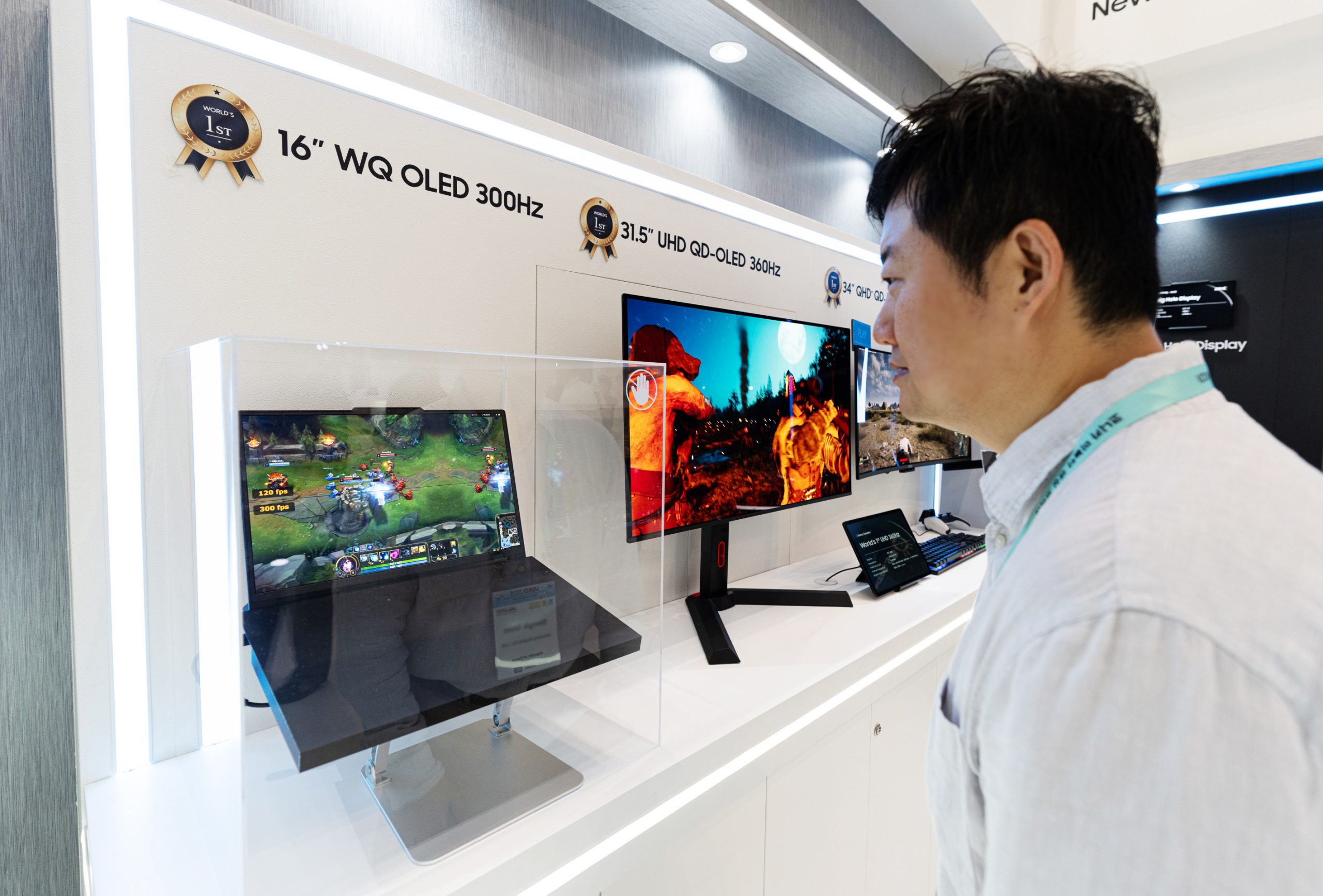 A display showcases a 16” WQ OLED 300Hz screen playing a video game, with a person observing alongside additional monitors labeled 31.5” UHD QD-OLED 360Hz and 34” GHD QD-OLED.