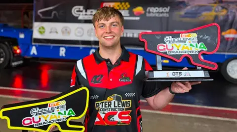 Bourne HPP A man in a red and black T-Shirt smiling at the camera holding two trophies in the shape of a race track.