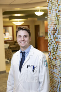 A man in a white lab coat stands indoors near a mosaic-tiled wall in a medical facility, smiling at the camera.