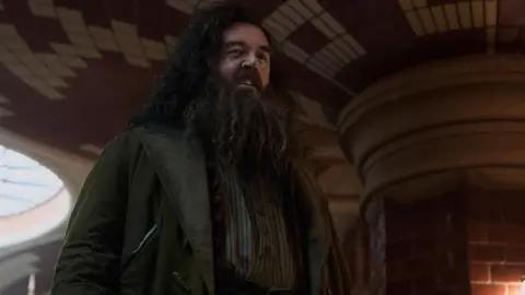 Aidan Monaghan/HBO Nick Frost in green jacket with long hair, moustache and beard
