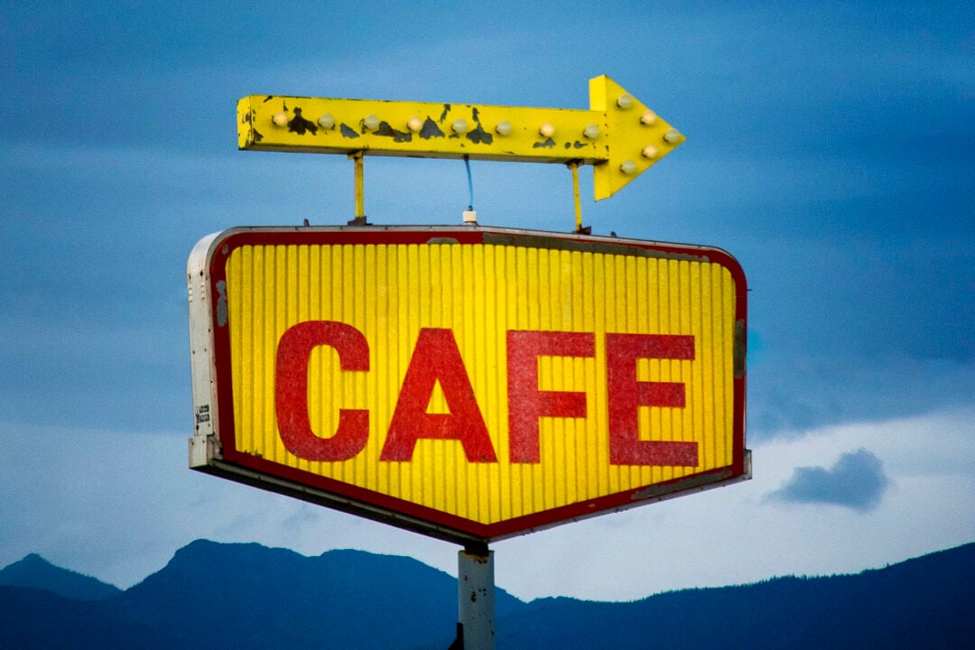A vintage sign with large red letters spelling "CAFE" on a yellow background is topped with a weathered, yellow arrow pointing right. The sky is overcast, and mountains are faintly visible in the background.