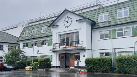 Howard Berry An Art Deco white building with a clock above the entrance which is on two floors with a balcony on the top floor. "Fairbanks" is written above the entrance on the ground floor. The building overall is on three floors and has a hedge and lamps outside it.