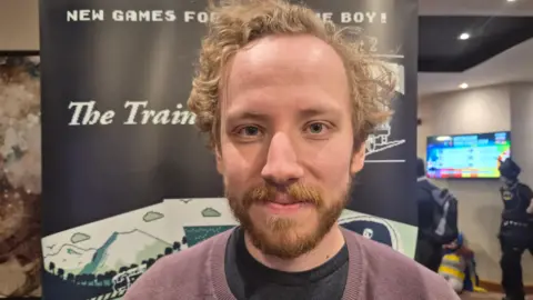 BBC A young man with curly hair and a beard wearing a purple jumper infront of a banner that says "new games for Game Boy!"