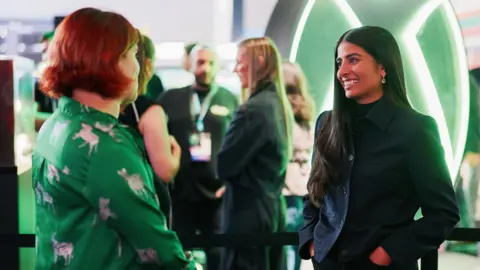 Marius Faulhaber Two women stand in a convention hall with a big green Xbox logo behind them. On the left, a person with a green shirt and short red hair. On the right, a woman with long brown hair and a dark jacket.