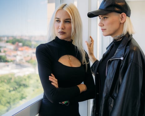 Liv and Mim stand by a window, one wearing a black leather cap and jacket