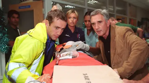 BBC A picture taken from the production of the television programme Holby City. It features male and female actors around a stretcher with a patient on it. There are two nurses in the background played by female actors and two male actors are in the foreground helping to push the stretcher.