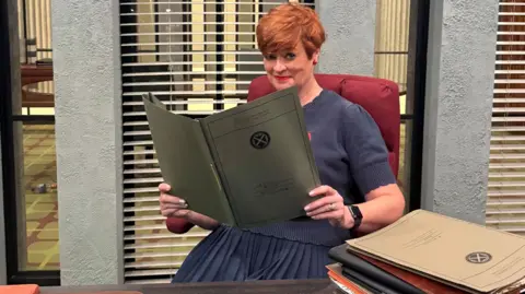 BBC Pauline McLean sitting behind a police desk holding a crime report. There are blinds on windows behind her with a room behind