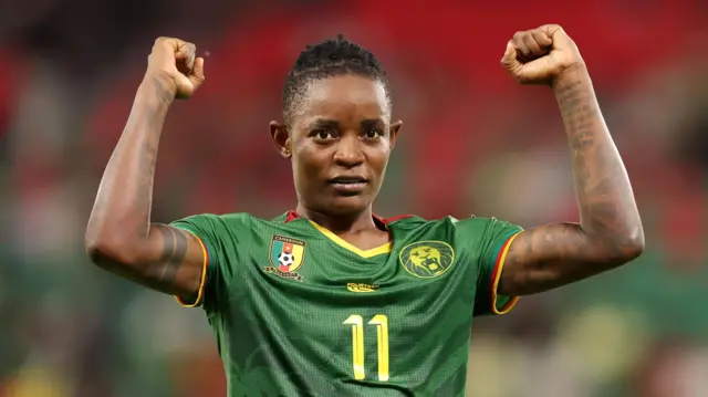 Marie Ngah Manga, wearing a green Cameroon shirt with a yellow number 11 on the chest, holds up both arms with fists clenched in celebration