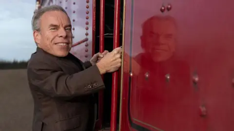 Aidan Monaghan/HBO Warwick Davis boarding a red steam train and smiling at the camera