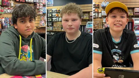 Darren Rozier/BBC A composite image of three boys smiling at the camera. They are sitting at the same table within a games cafe. The boy on the left has brown hair and wears a green hoodie. The boy in the middle has blond curly hair and is wearing a black T-shirt with a chain on. The boy on the right wears a yellow cap and a black T-shirt.
