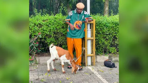 Courtesy of Promise Okoro Promise stands by a lamp post in orange trousers and a green top. The goat is next to him tethered to the lamp post.