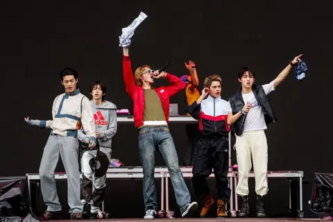 FilmMagic via Getty Images Five boyband members wearing jackets and jeans performing on a stage