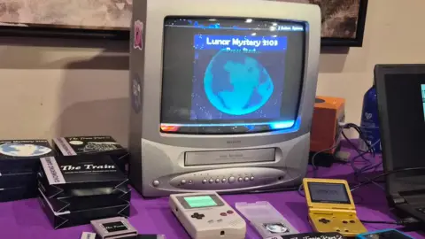 A small old grey television with the words Lunar Mystery 2103 on the screen and an image of the moon sits on a table covered in a bright purple cloth. The television is surrounded by other types of retro technology such as game cartridges and different handheld Game Boy consoles.
