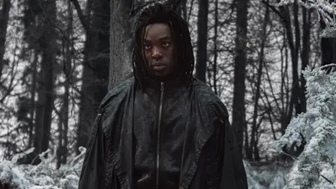 Lara Cornell/HBO Paapa Essiedu in a black zip-up jacket and cape looking moody in a snowy forest