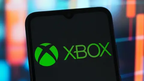 Getty Images Xbox logo displayed on a smartphone screen, in front of a backdrop of a blurred financial chart with red candlestick indicators.