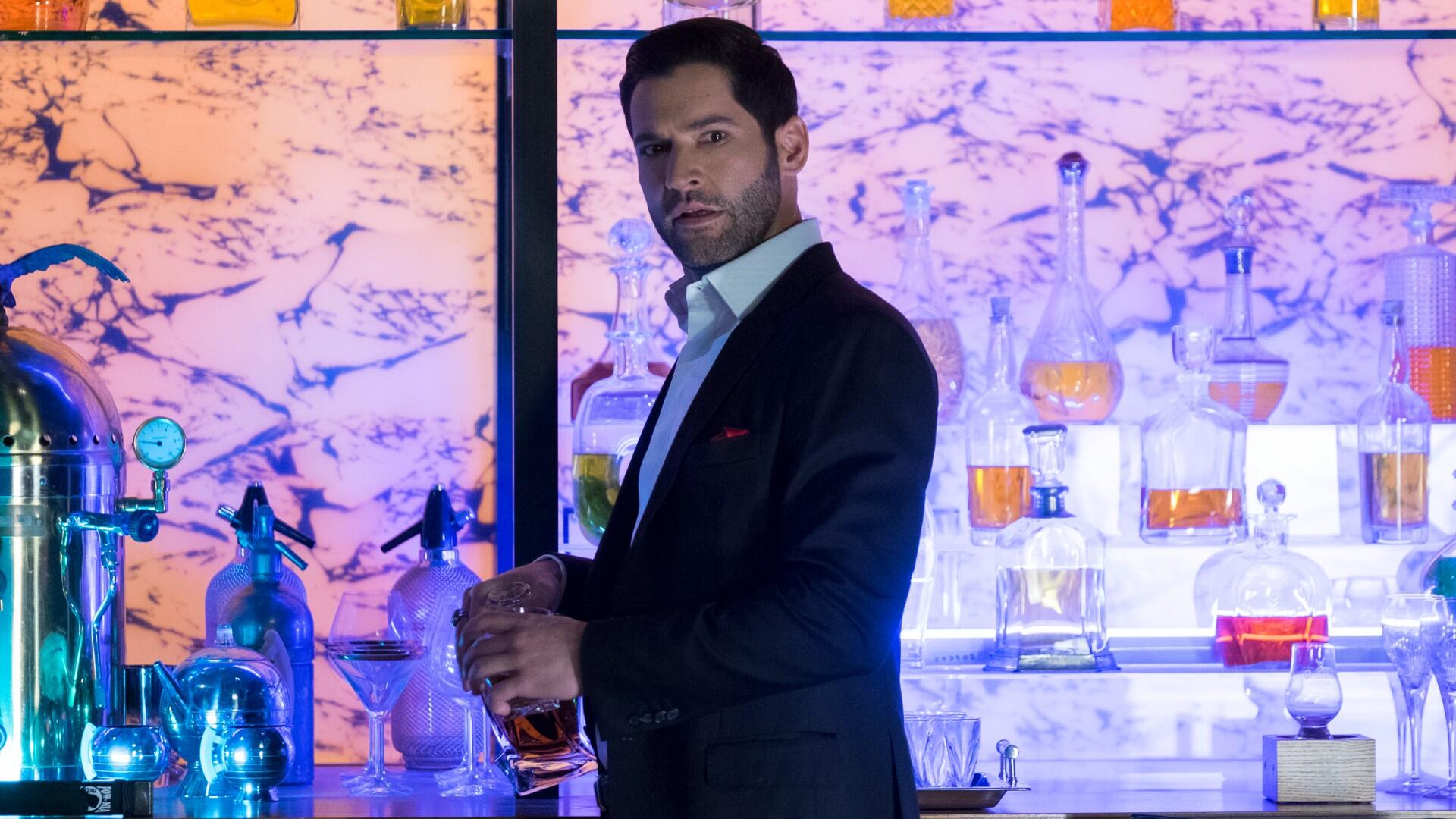 A bearded man in a suit stands at a bar with bottles of different colored liquid.