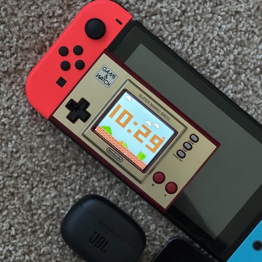 A Nintendo Game & Watch: Super Mario Bros. displaying the time sits on top of a Nintendo Switch with red Joy-Con controllers. A black JBL earbud case is nearby on a carpeted surface.
